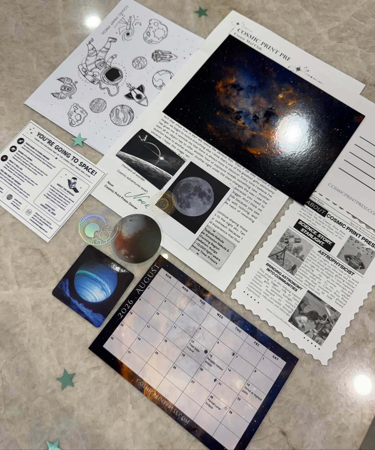 A space-themed educational activity kit laid out on a beige marble surface, featuring printed cards, a moon phase tracker grid, a small galaxy card, a sticker, and printed sheets with black-and-white space illustrations and a dark space scene photo. Photos are taken by Noor and Astrophysicist who uses her telescope to capture the images featured in her astronomy space themed mail club