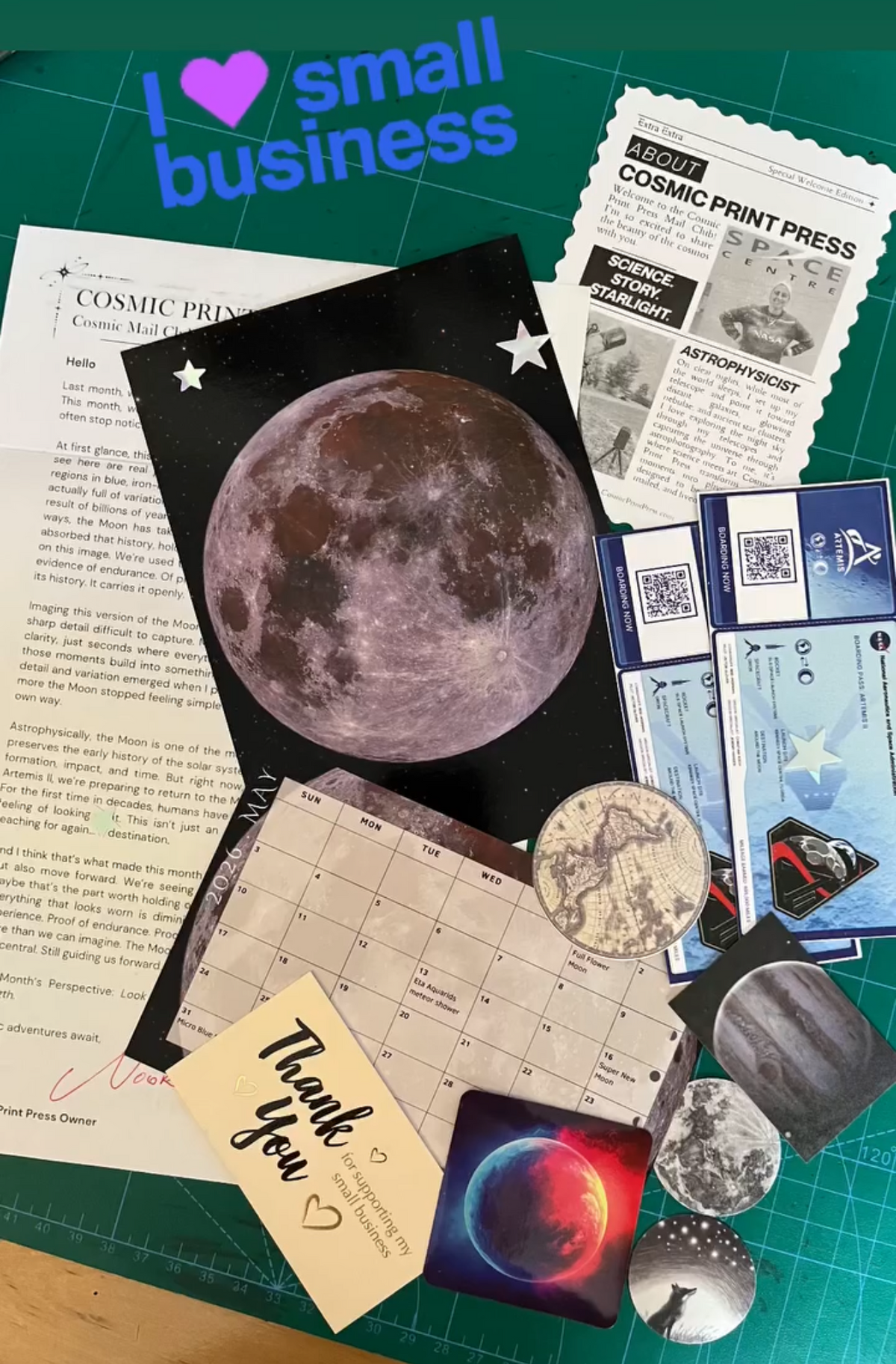 A flat-lay display of various small business themed items, including a moon print, a 'Thank you' card, a 'Cosmic Print Press' coupon, and several small printed cards and stickers, arranged on a green cutting mat. Photos are taken by Noor and Astrophysicist who uses her telescope to capture the images featured in her astronomy space themed mail club