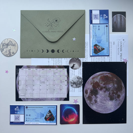 A flat lay of a moon-themed stationery set on a light background, including an olive green envelope with moon phase graphics, printed cards, a calendar page, a large moon print, and small decorative elements. Photos are taken by Noor and Astrophysicist who uses her telescope to capture the images featured in her astronomy space themed mail club Cosmic print press 